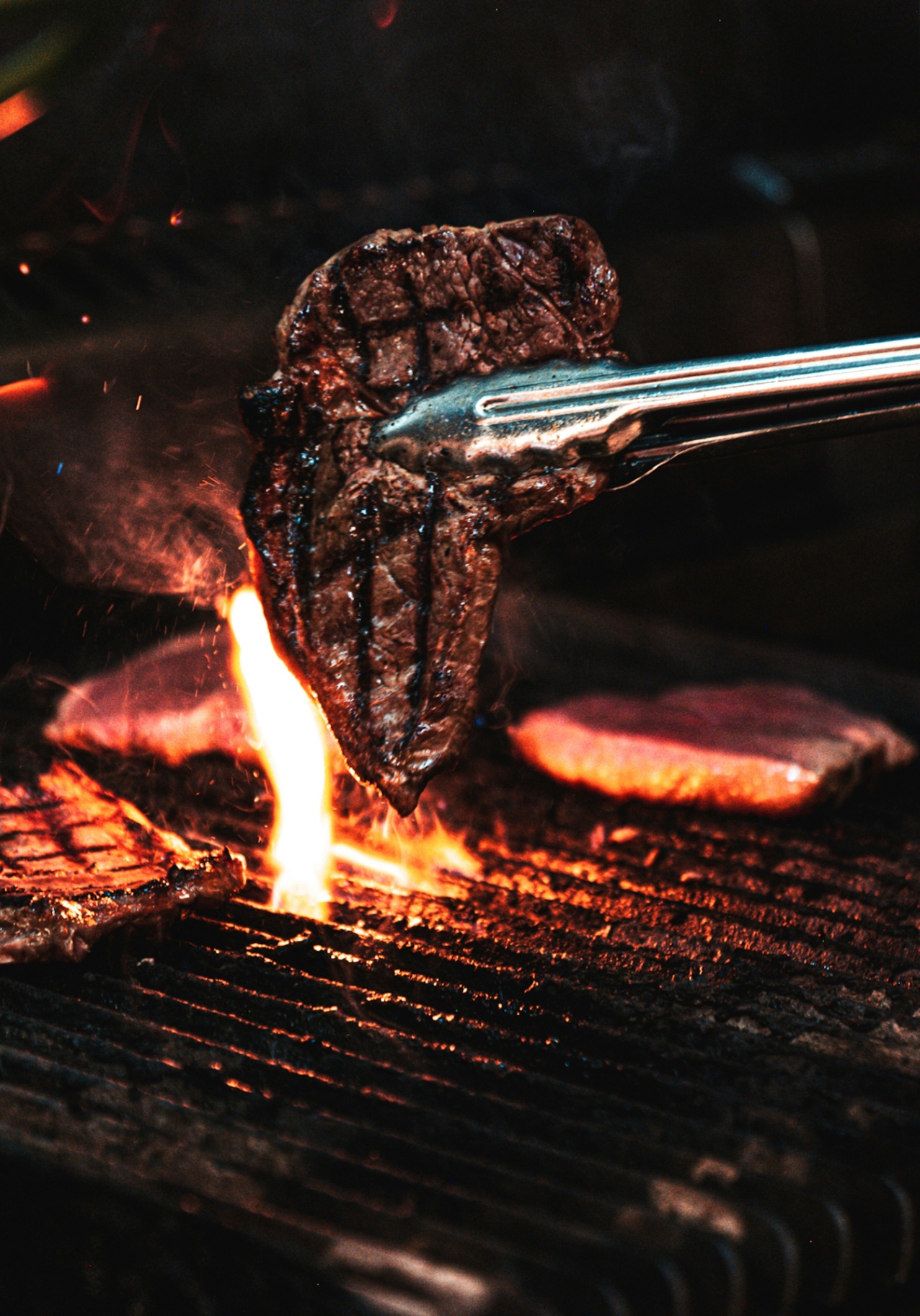 Pitmaster lifting a seared steak off the live-fire grill with tongs
